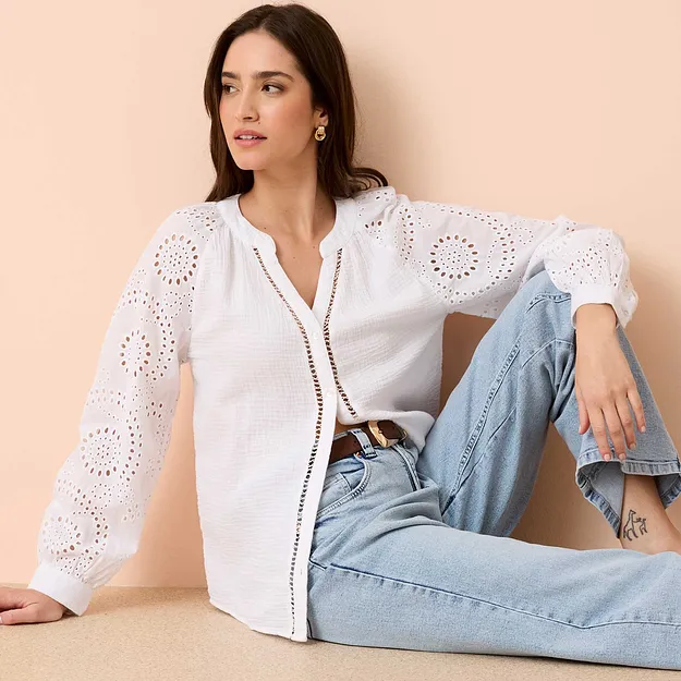 Camicetta bianca con ricami traforati e maniche lunghe, abbinata a jeans azzurri e cintura.