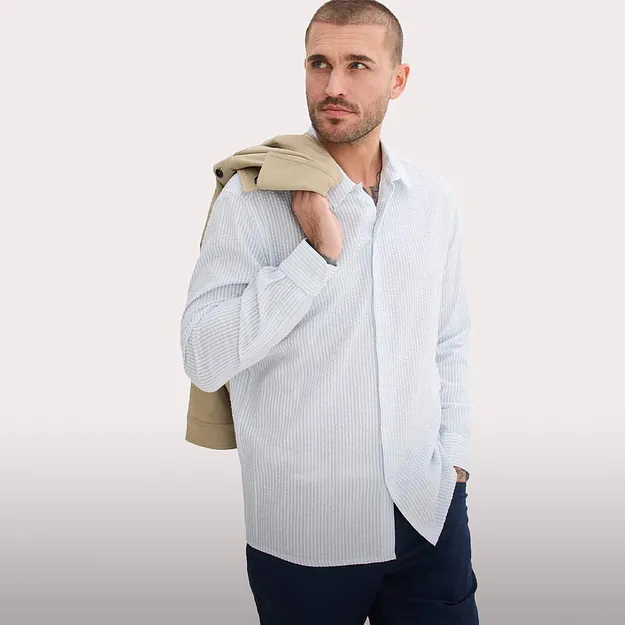 Camicia a righe azzurra, pantaloni scuri e un maglione beige appoggiato sulle spalle.