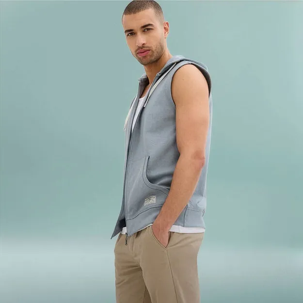 Un uomo indossa un gilet con cappuccio azzurro sopra una maglietta bianca e pantaloni beige.