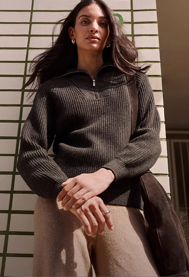 Maglione scuro a coste con cerniera, pantaloni chiari e borsa in pelle scamosciata marrone.