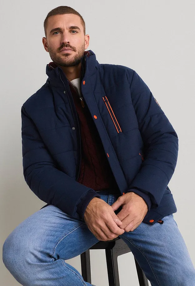 Giacca trapuntata blu scuro con dettagli arancioni, maglione rosso lavorato a maglia e jeans chiari.