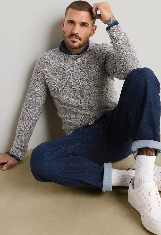 Maglione grigio lavorato a maglia sopra una camicia blu, jeans scuri, calzini bianchi e scarpe da ginnastica.