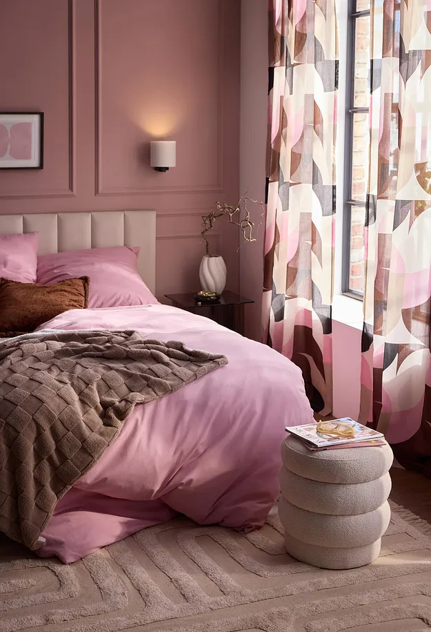 Biancheria da letto rosa, una coperta marrone, tende a motivi e un tappeto soffice.