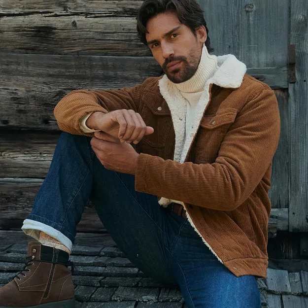 Uomo con giacca di velluto a coste marrone, maglione a collo alto bianco, jeans e stivali pesanti.