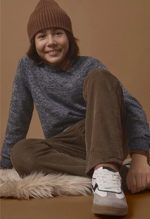 Un ragazzo indossa un berretto marrone, un maglione blu, pantaloni marroni e sneaker.