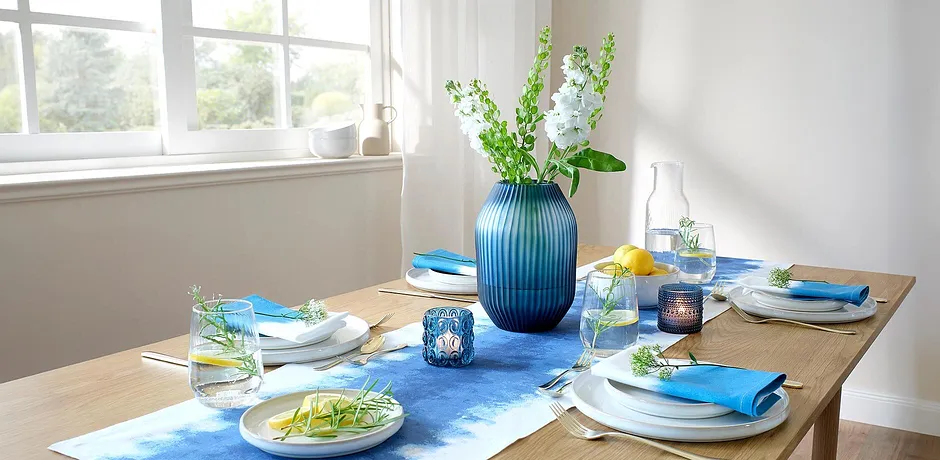 Un runner da tavolo blu decora il tavolo da pranzo, in contrasto con piatti bianchi e accessori colorati.