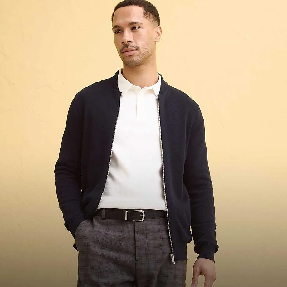 Cardigan scuro sopra una polo bianca, abbinato a pantaloni grigi.