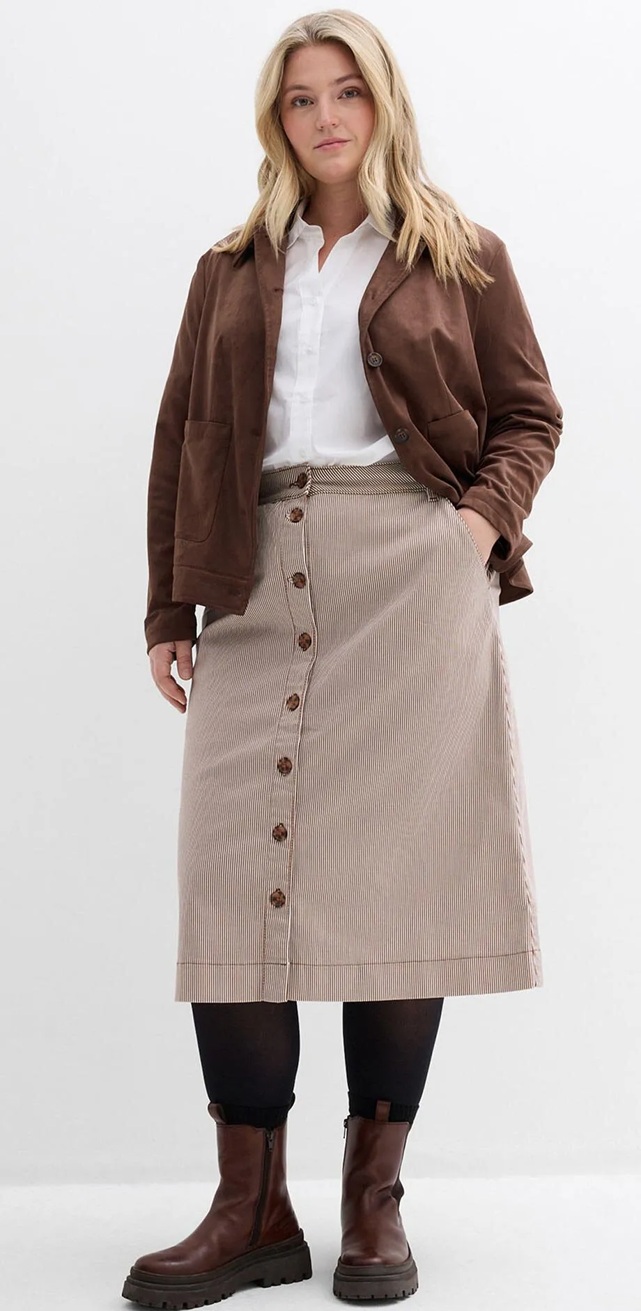 Giacca marrone, camicetta bianca, gonna a righe con bottoni e stivali marroni.