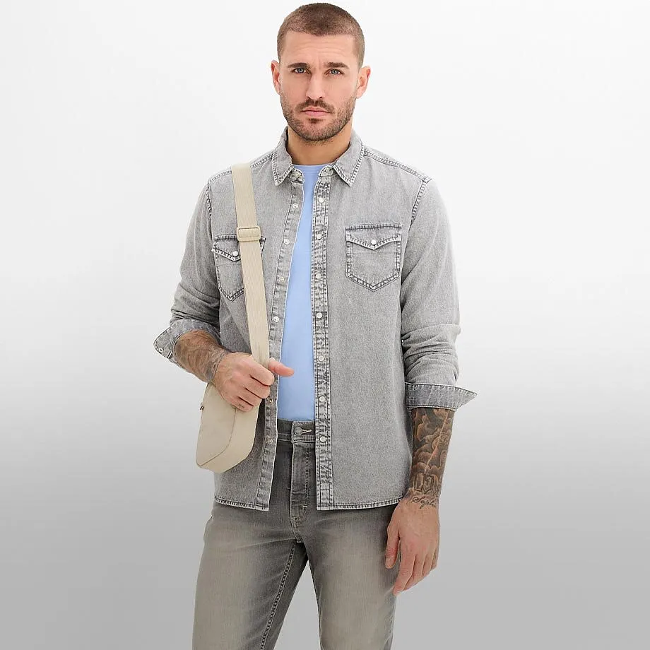 Camicia di jeans chiara sopra una maglietta azzurra, abbinata a jeans grigi e una borsa a tracolla beige.