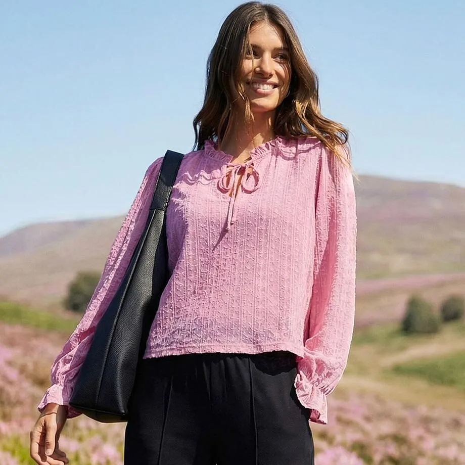 Maglia rosa a maniche lunghe con laccetto allo scollo, maniche ampie e pantaloni neri.