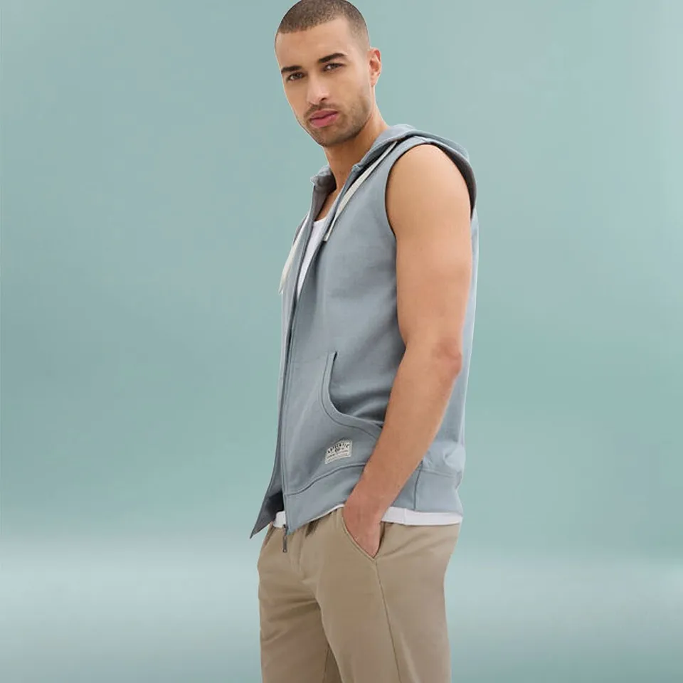 Un uomo indossa un gilet in felpa con cappuccio azzurro sopra una maglietta bianca e pantaloni beige.