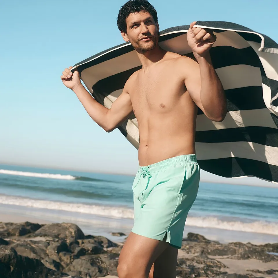 Un uomo indossa un costume da bagno verde menta e tiene in mano un asciugamano a righe sulla spiaggia.