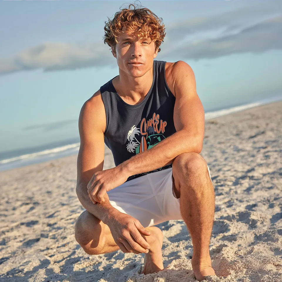 Un uomo indossa una canottiera blu con una stampa e dei pantaloncini bianchi in spiaggia.