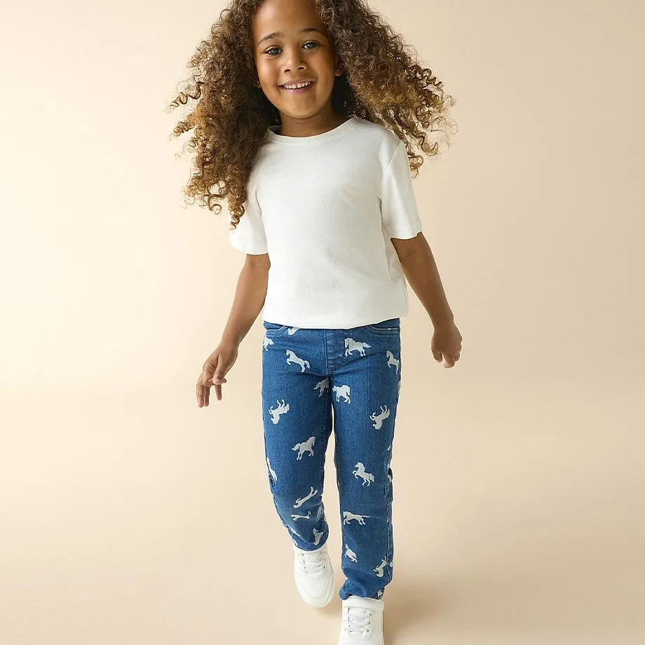 Una bambina indossa una maglietta bianca, jeans blu con un motivo a cavalli e sneaker bianche.
