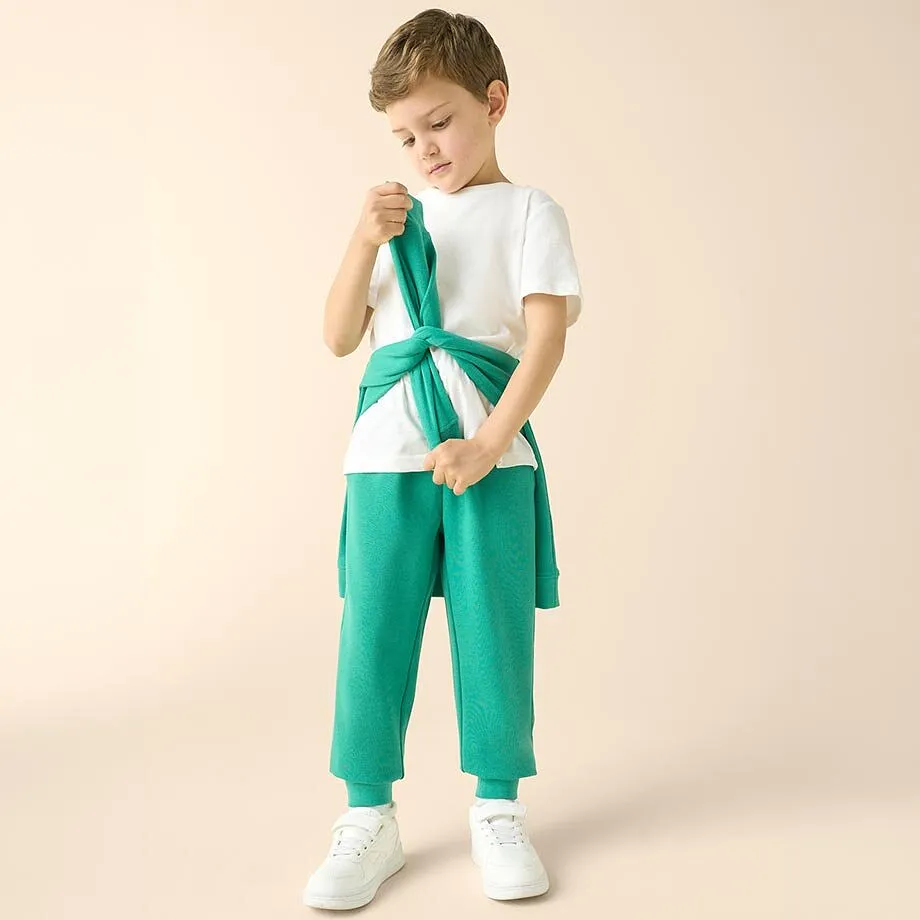 Bambino con maglietta bianca, pantaloni verdi e felpa annodata.