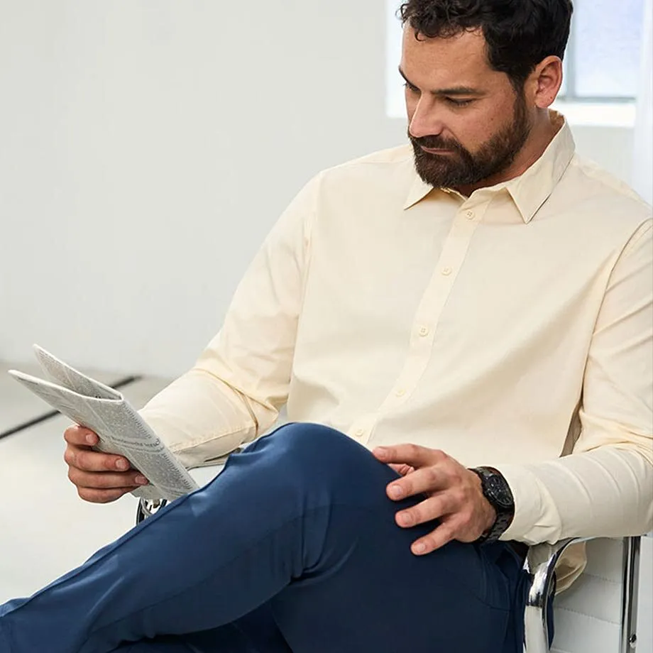 Un uomo indossa una camicia beige, pantaloni blu scuro e un orologio da polso nero.