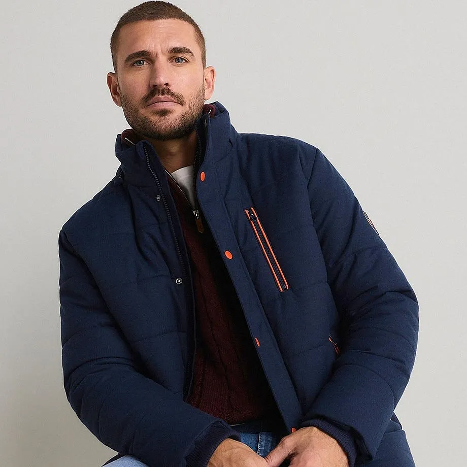 Un uomo indossa una giacca trapuntata blu scuro, un maglione bordeaux e dei jeans azzurri.