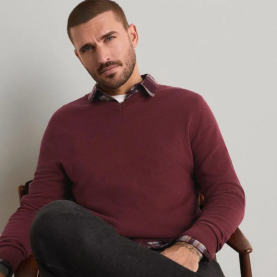 Un uomo indossa un maglione bordeaux, una camicia a quadri e dei jeans scuri.