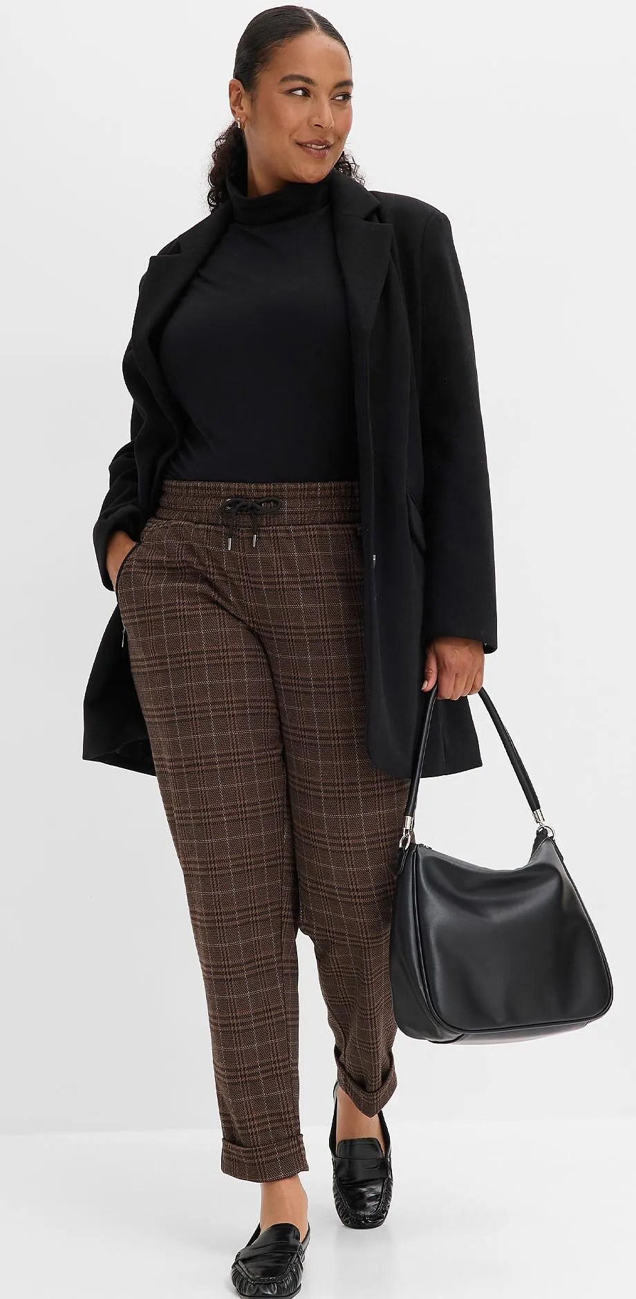 Maglione a collo alto nero, blazer lungo, pantaloni a quadri, scarpe eleganti e borsa nera.