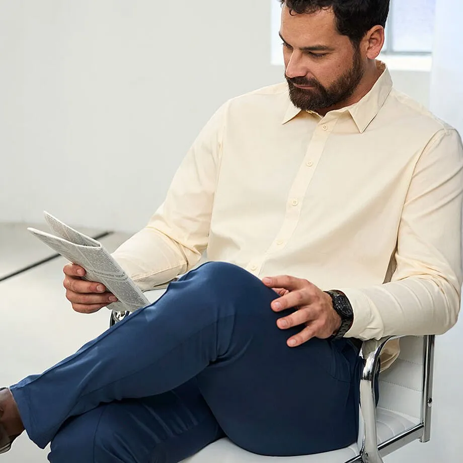 Un uomo indossa una camicia beige, pantaloni blu scuro e un orologio da polso nero.