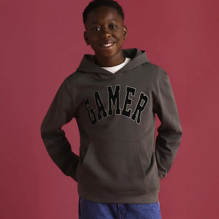 Felpa con cappuccio grigia e nera con la scritta &ldquo;Gamer&rdquo;, abbinata a jeans blu.