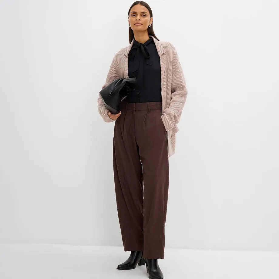 Indossa una camicetta nera, un cardigan di maglia beige, pantaloni marroni e stivaletti neri.