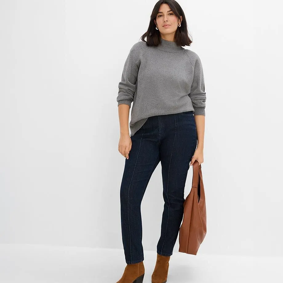 Maglione grigio con collo alto, jeans scuri con cuciture, stivaletti marroni e borsa.
