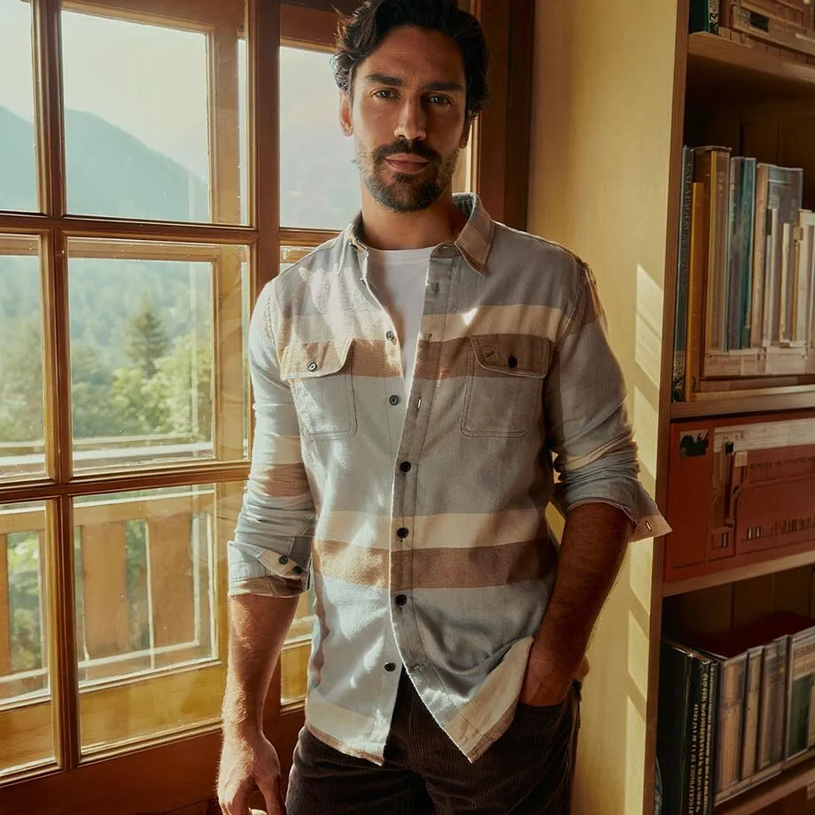 Indossa una camicia a righe sopra una maglietta bianca e dei pantaloni scuri.