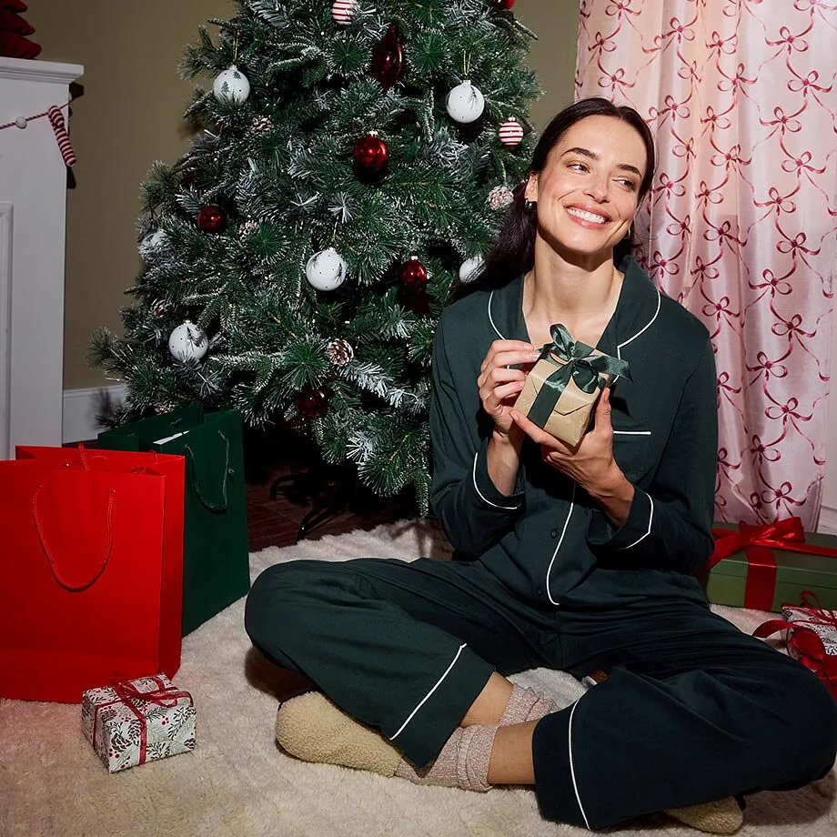 Donna seduta davanti a un albero di Natale addobbato con pigiama verde bosco con riga bianca.