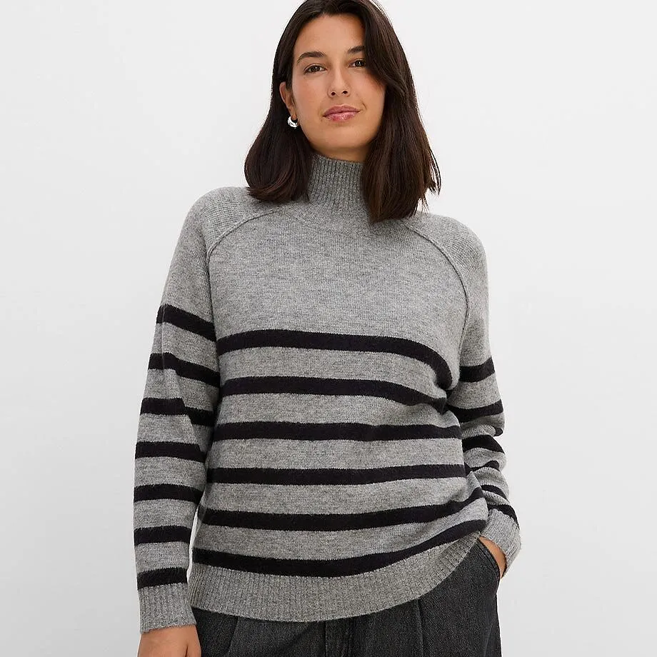 Maglione a collo alto lavorato a maglia, grigio con strisce trasversali nere, abbinato a pantaloni scuri.